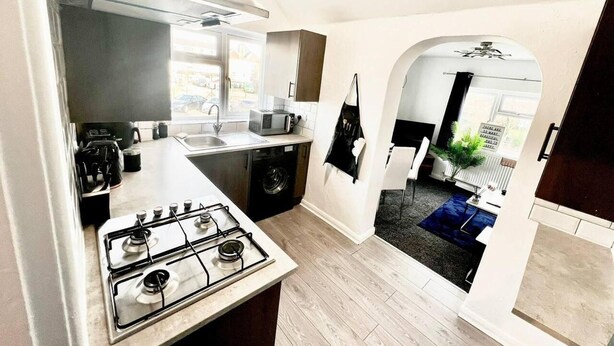 A compact kitchen with a gas stove, a microwave, and a sink. There is a small dining area with a table and chairs, and a view of the outside through an archway. - Beautiful 2-bed House in Watford (Watford)