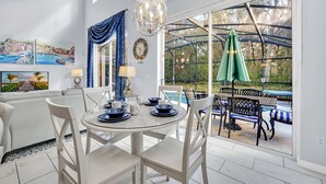 A dining area with a glass table, white chairs, and a view of a swimming pool through large windows. - Stylish 6 bedroom! Near Disney! with private pool! (Kissimmee)