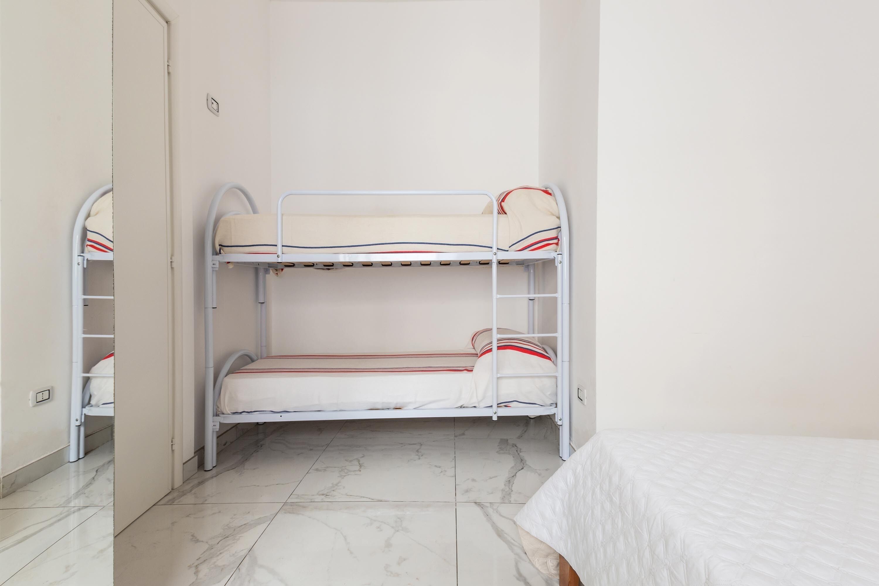 A bunk bed with white bedding and red stripes, a marble-tiled floor, and a plain wall.