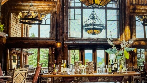 Un intérieur rustique en bois, agrémenté de grandes fenêtres, d’un lustre et d’une table à manger dressée avec des verres et des bouteilles.