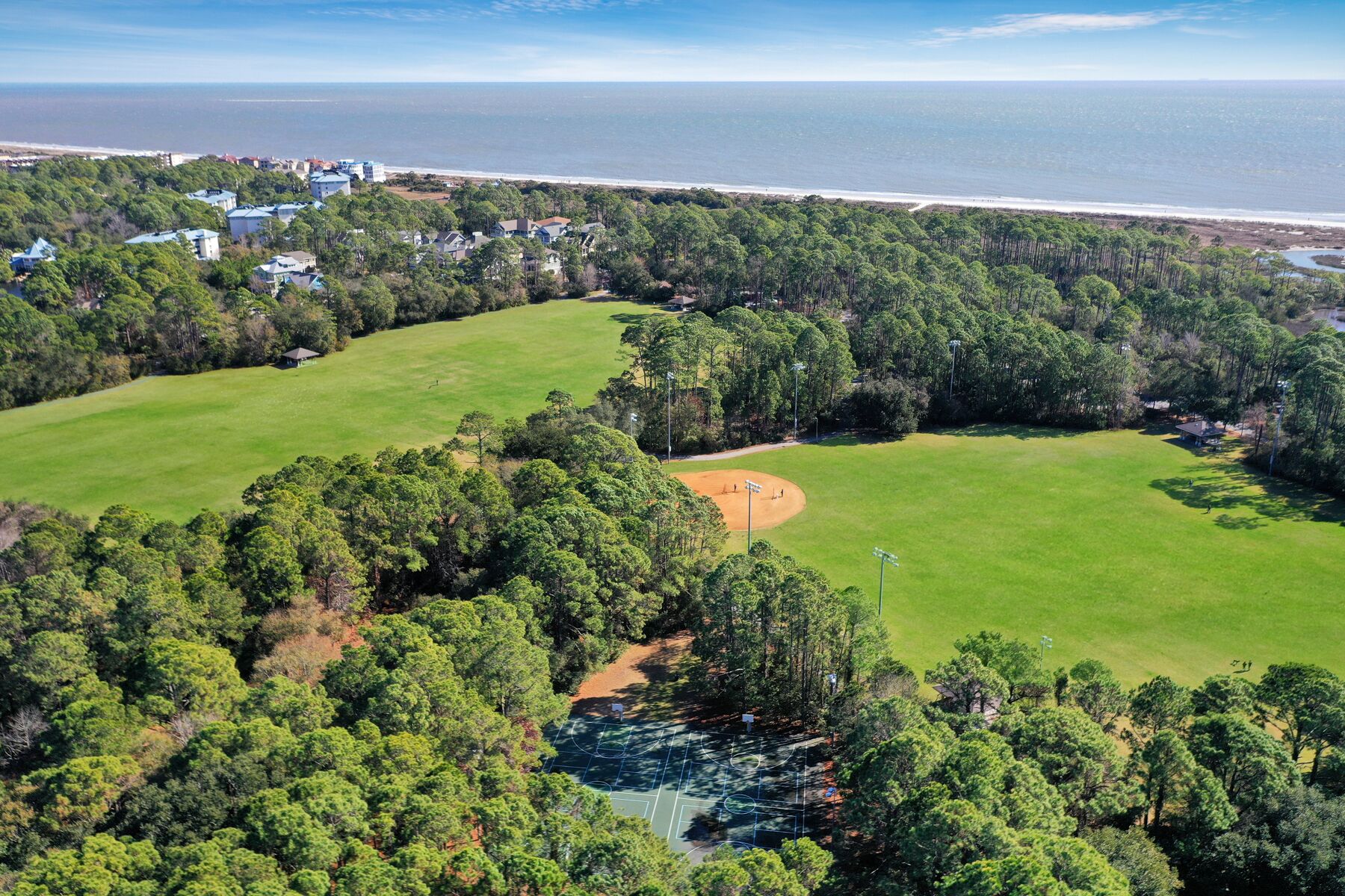 Vista aérea de un campo de golf con varias zonas verdes y un diamante de béisbol, rodeado de un bosque denso y una playa al fondo.