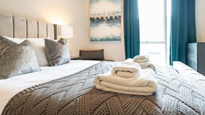 A neatly made bed with a patterned blanket, a stack of folded towels, and a painting on the wall.