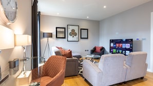 A modern living room with a sofa, armchairs, a coffee table, a clock, framed artwork, and a television.