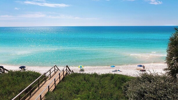 A beach with a wooden walkway leading to the shore, clear blue water, and a few umbrellas.
