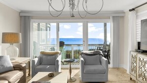 A modern living room with a chandelier, two armchairs, a sofa, a coffee table, and a TV. The room has large windows with a view of the ocean.