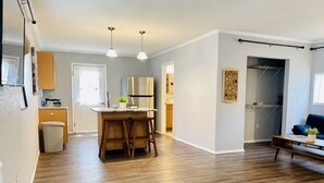 A spacious living area with a wooden floor, a kitchen with a refrigerator, a dining table, and a bar stool. There is a coat rack and a bench in the corner. - Farm living by Santos Trails & the Villages! (Summerfield)