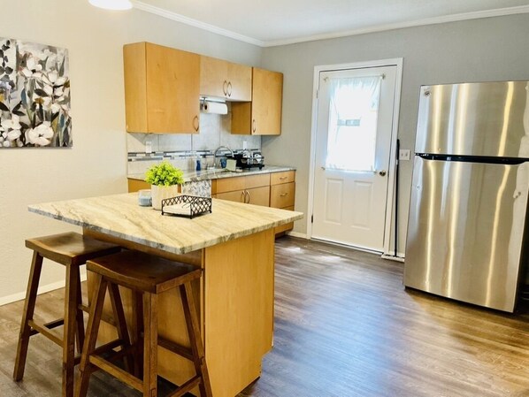 A kitchen with wooden cabinets, a marble countertop, a stainless steel refrigerator, and a door with a window.