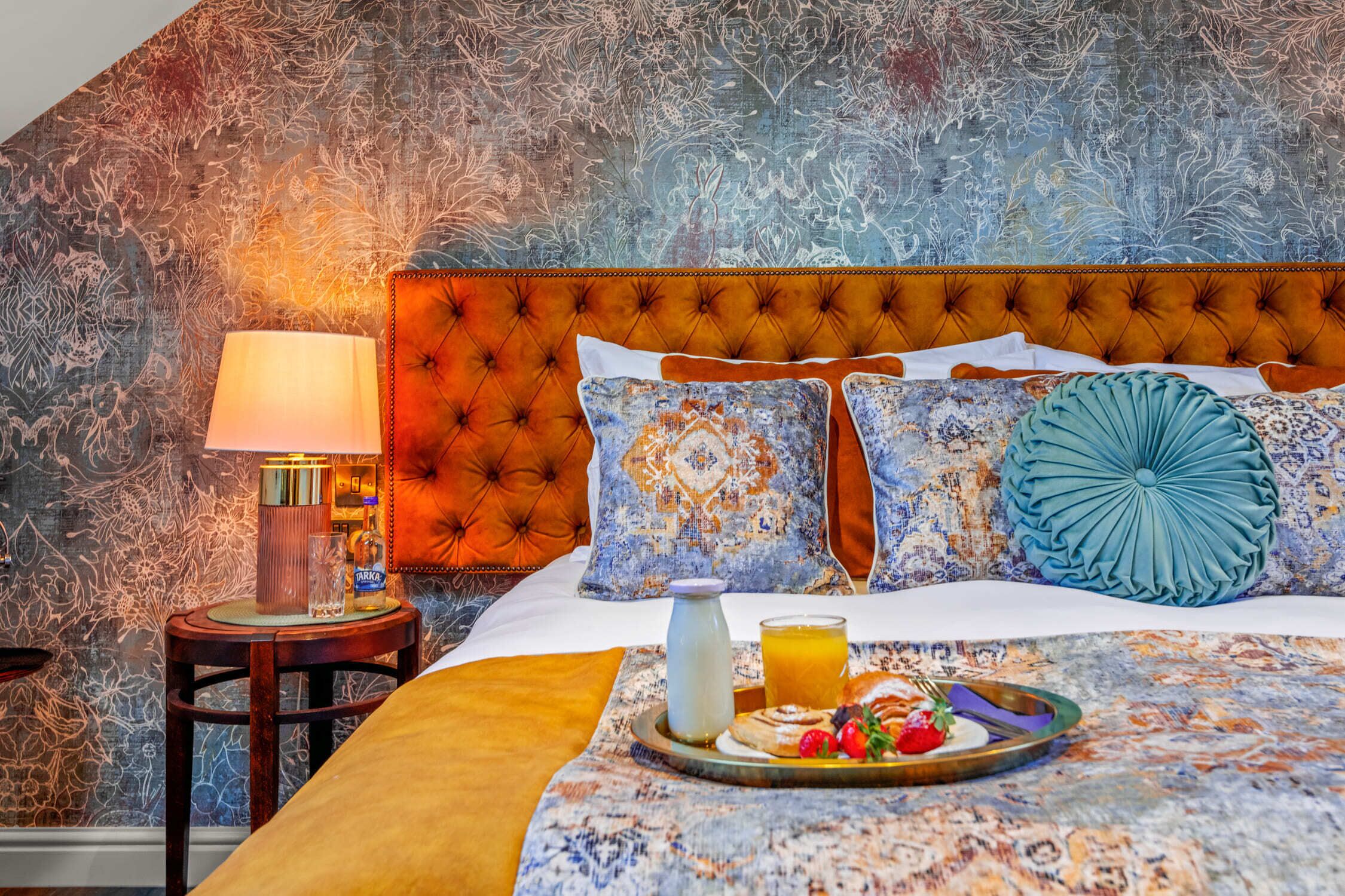 Habitación de hotel con cabecera capitonada, pared estampada y bandeja de desayuno con pastelería y jugo.