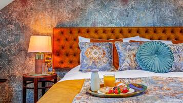 Habitación de hotel con cabecera capitonada, pared estampada y bandeja de desayuno con pastelería y jugo.