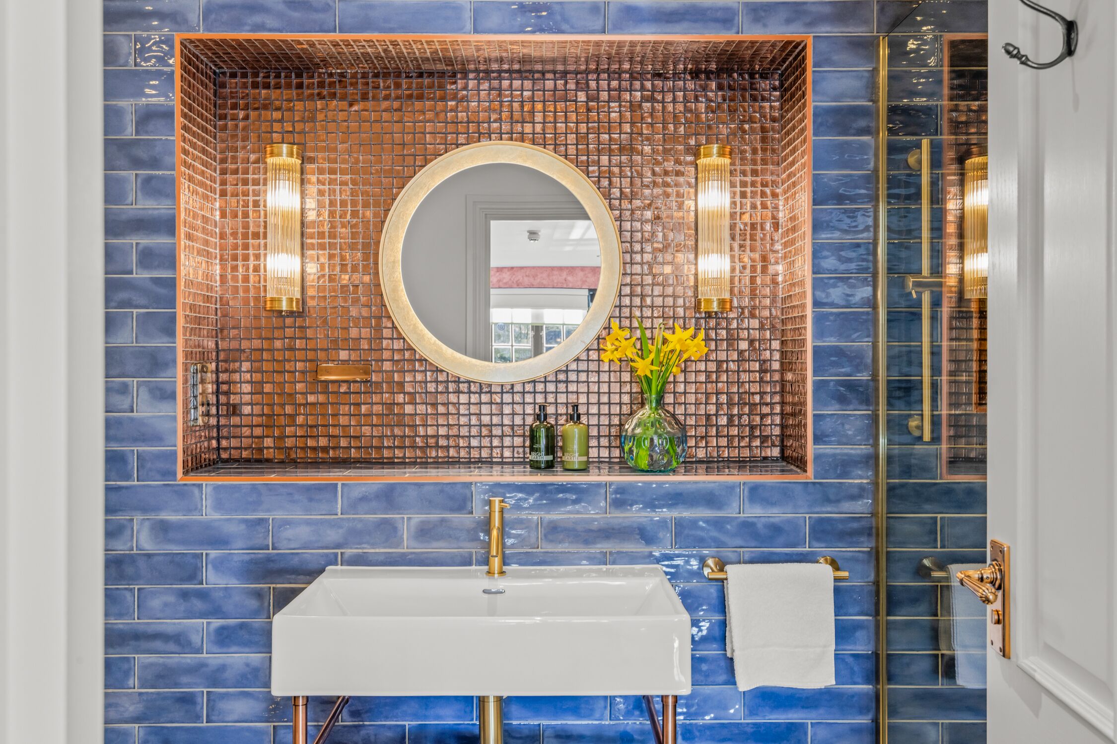 Un bagno con parete piastrellata di blu, un lavandino bianco, un rubinetto dorato, uno specchio rotondo e un vaso con fiori gialli.