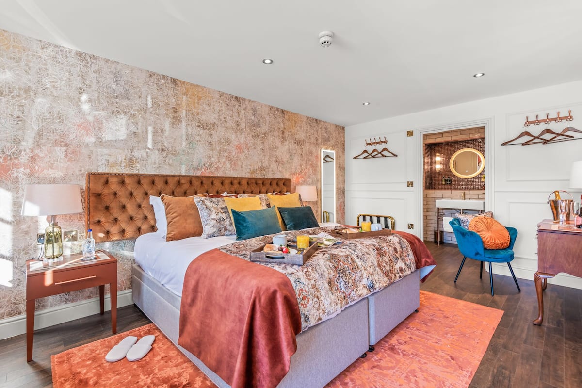 Quarto de hotel moderno com uma cama grande, um espaço de estar com uma poltrona, uma mesinha e um espelho.