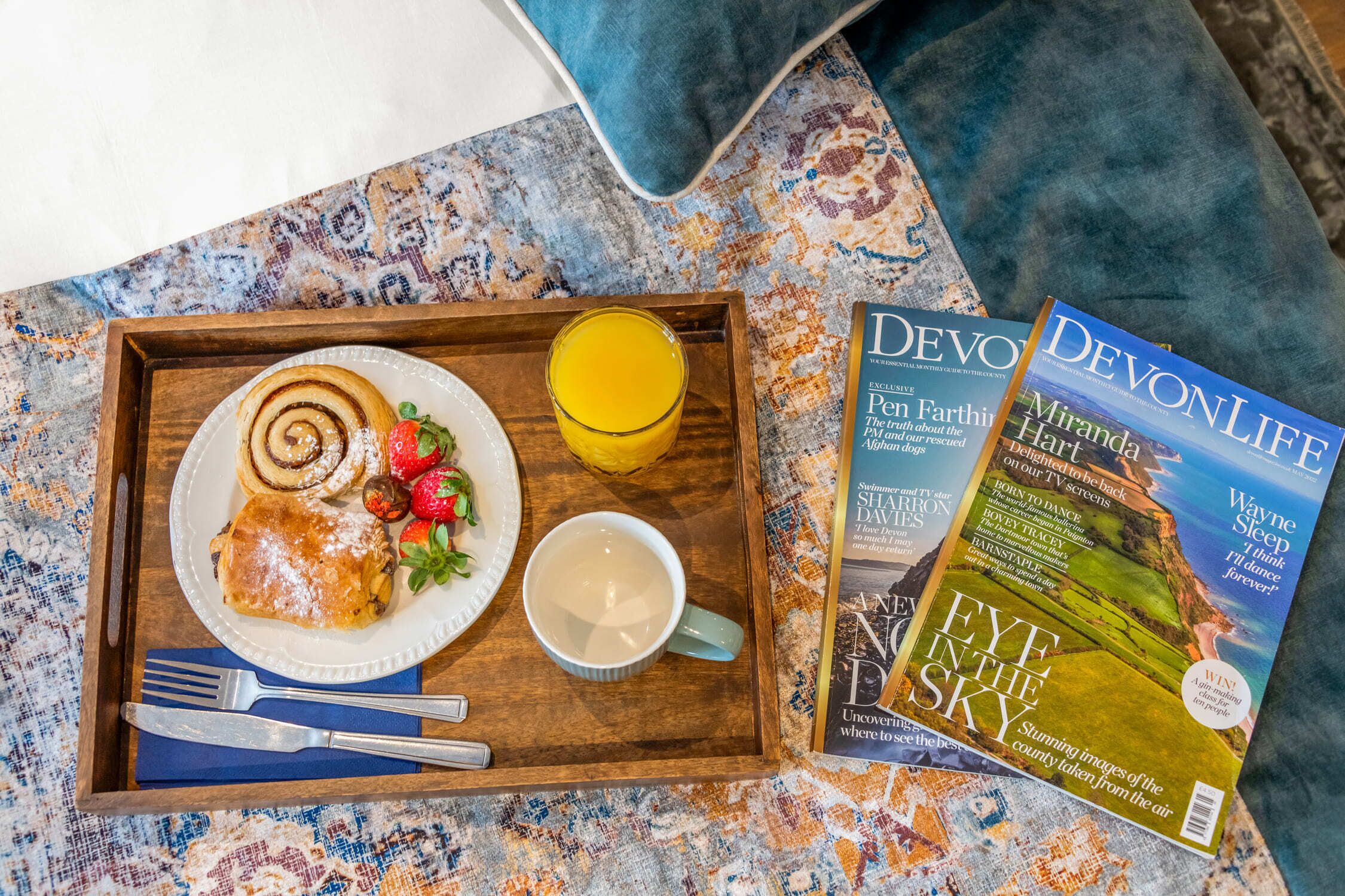 Una bandeja con un plato de comida, un vaso de zumo de naranja y una taza, junto con dos revistas sobre un mantel estampado.