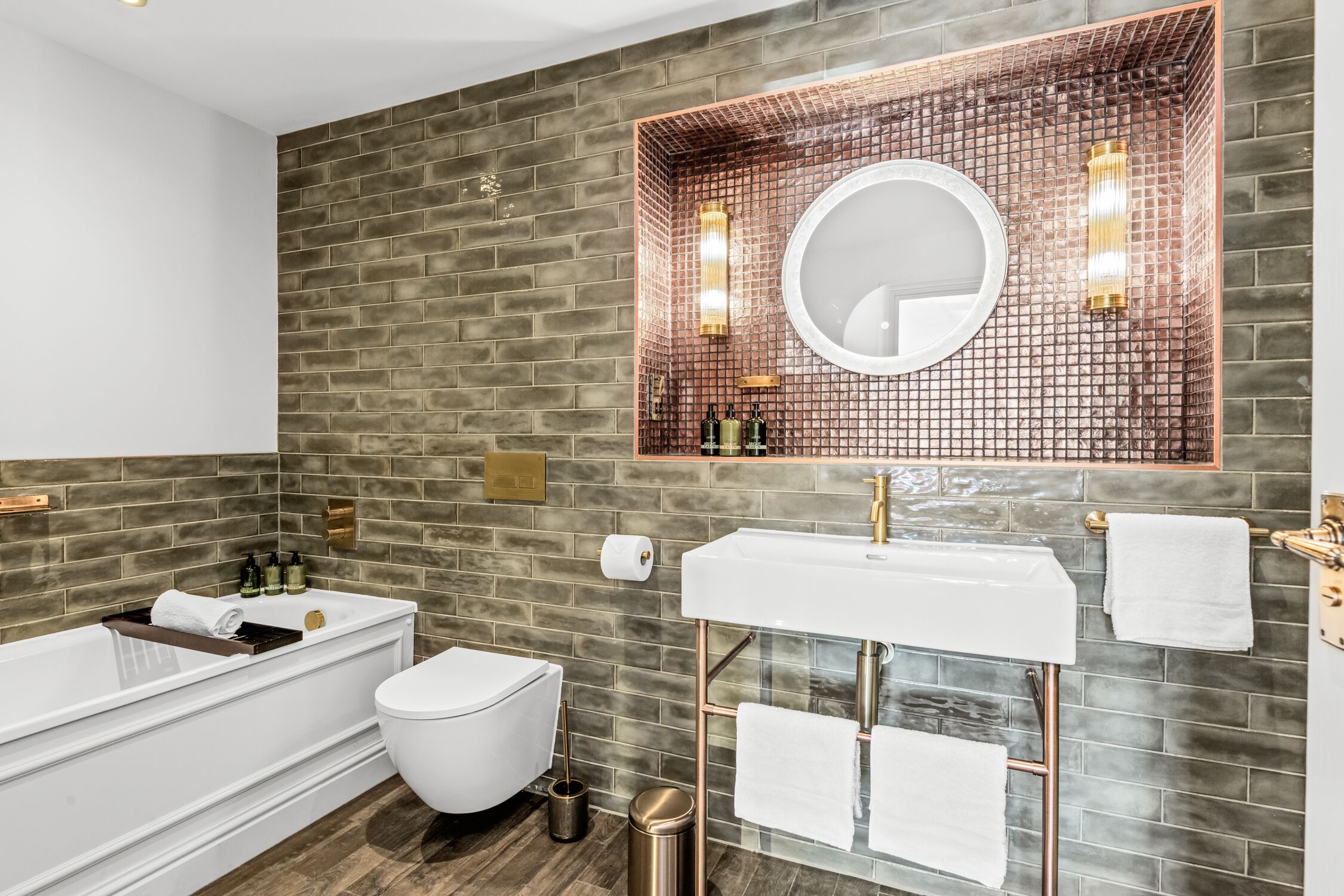 Un baño moderno con pared de ladrillo, una bañera blanca, un inodoro blanco y un lavamanos blanco con grifo dorado.