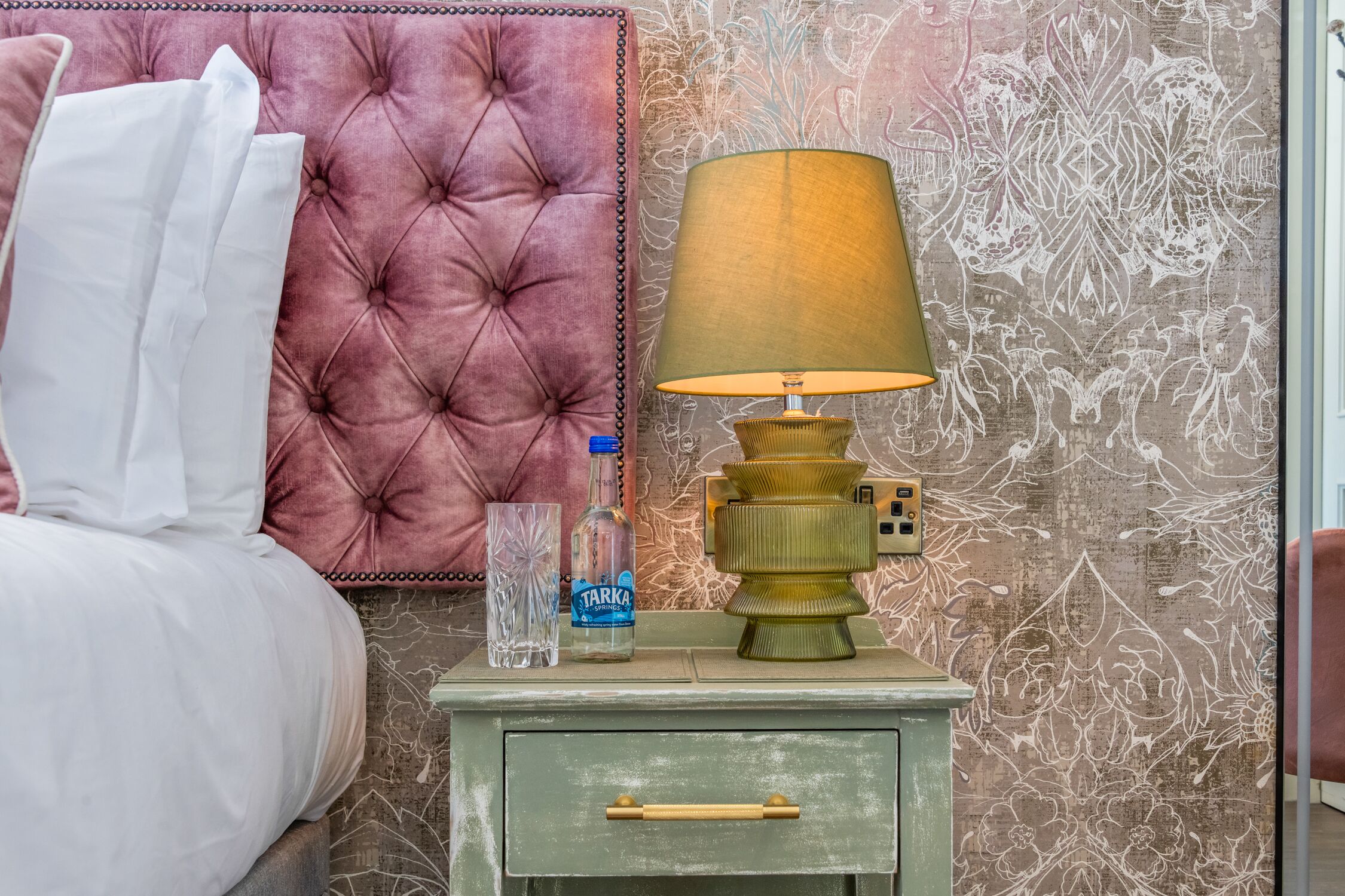 Un lit bien fait, avec du linge de lit blanc, une tête de lit rose à motifs, une table de chevet verte avec une lampe, un verre transparent et une bouteille d’eau.