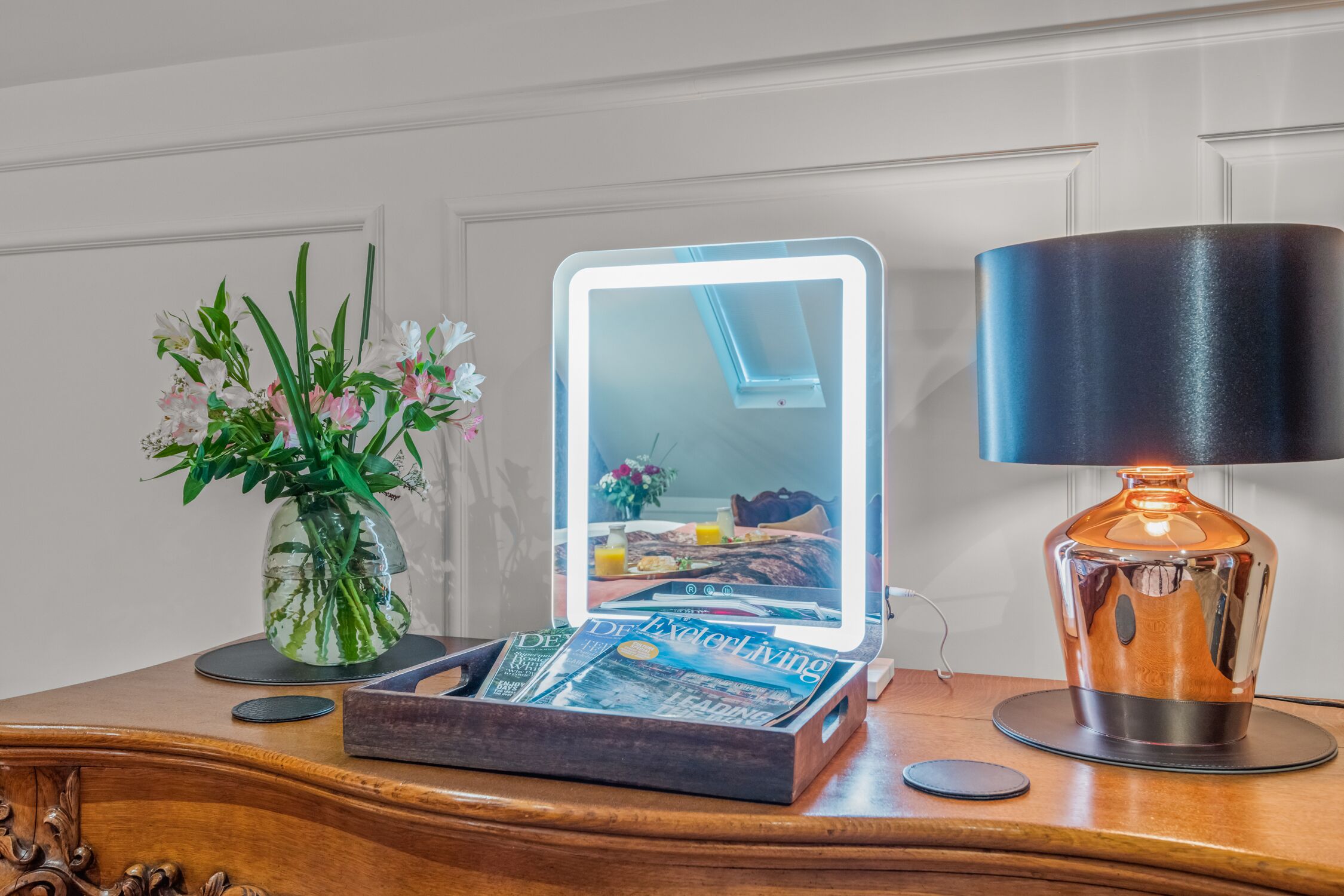 A wooden table with a magazine, a vase with flowers, a mirror with a light, and a lamp.