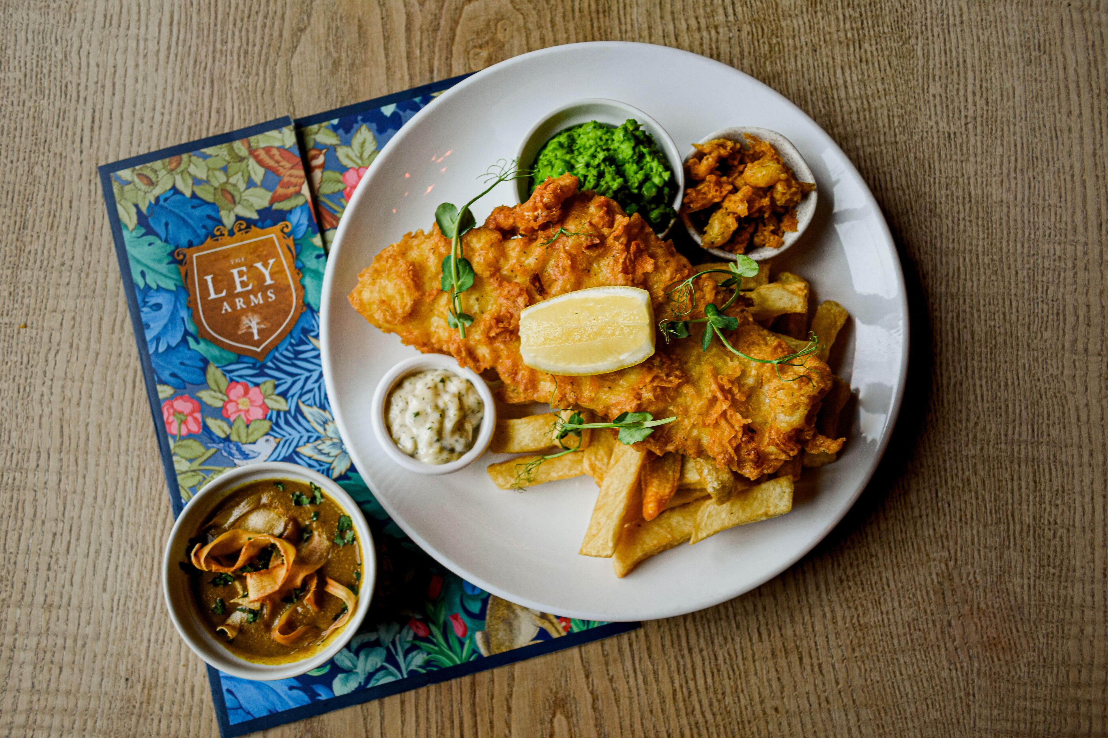 En tallrik med fisk och pommes frites, serverad med citron, tartarsås och mosade ärtor, på ett bord med en bordsduk med blomstermönster.