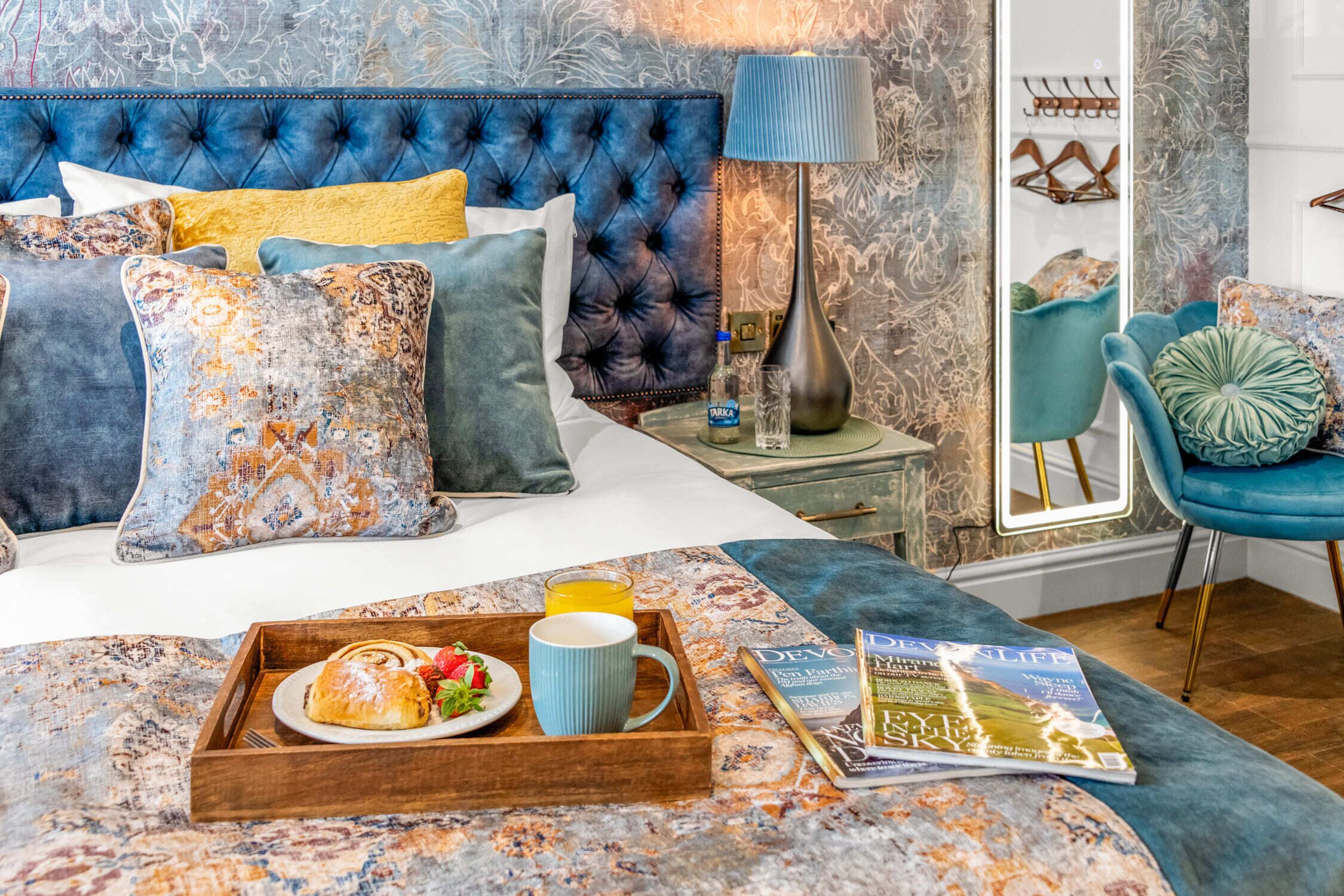 A bedroom with a tufted headboard, a bedside table with a lamp, a chair, and a tray with breakfast.
