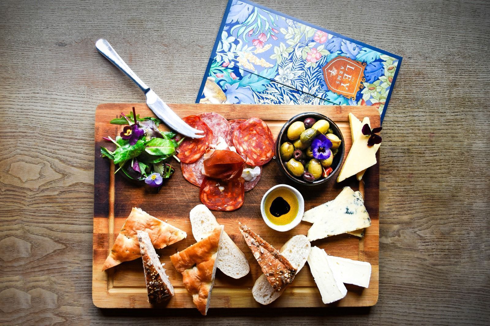 A wooden board with various cheeses, cured meats, olives, and bread, accompanied by a knife and a decorative card.
