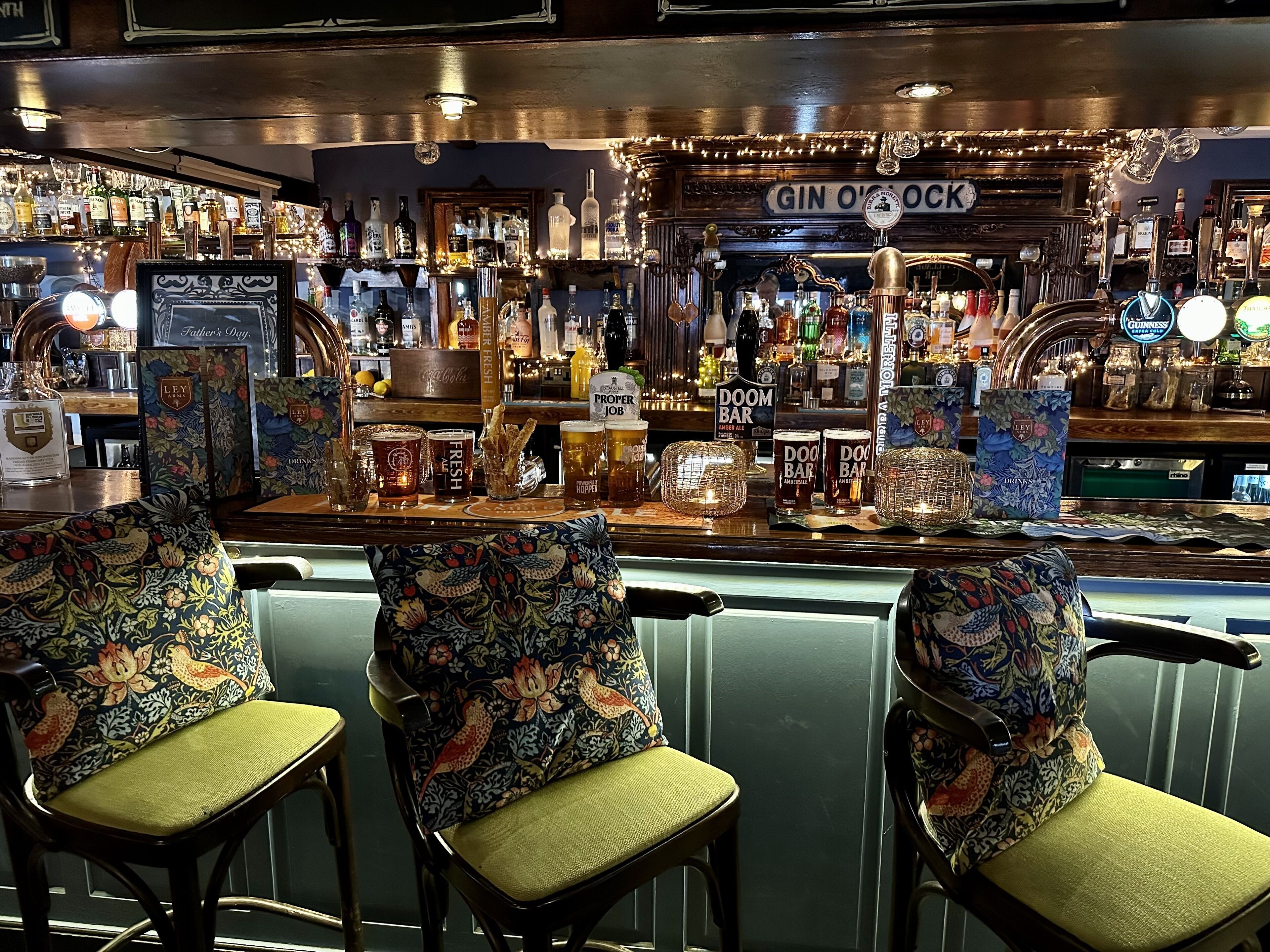 A bar with a variety of liquor bottles, a sign reading "GIN O'CLOCK", and two chairs with floral upholstery.