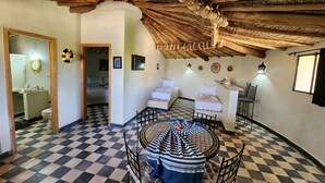 Une pièce avec un sol à carreaux, une table ronde entourée de chaises, un lit avec une literie blanche et un plafond en bois rustique.