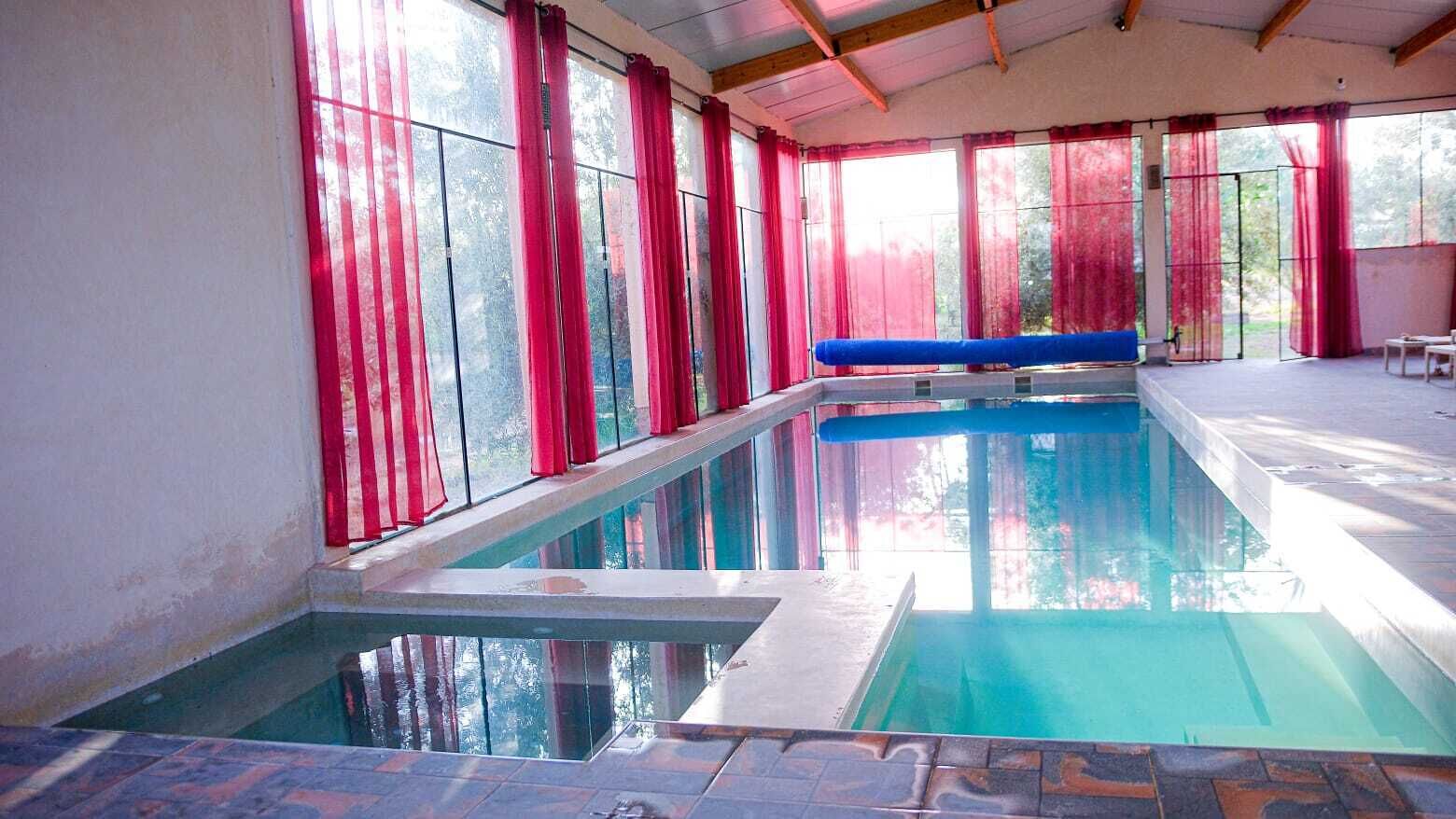 Una piscina interior con paredes de vidrio transparente, cortinas rojas y un banco azul.