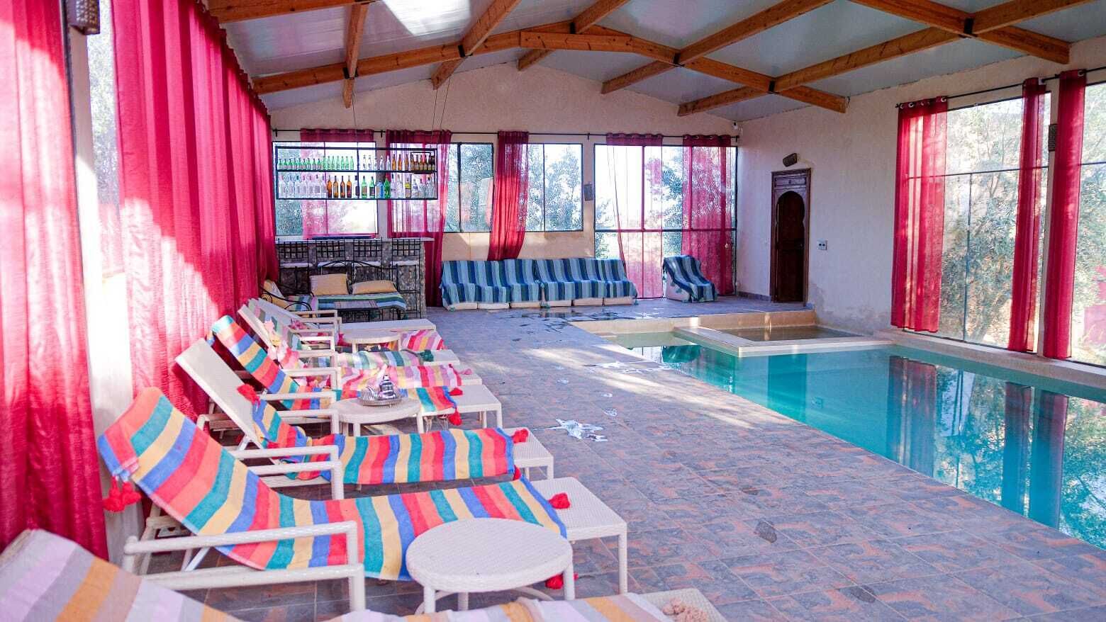 A sunlit indoor swimming pool area with lounge chairs and a seating area, surrounded by large windows and red curtains.