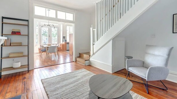 A modern living room with hardwood flooring, a staircase, a grey armchair, and a bookshelf. - Hall Street Inn, Relaxing | Historic | Fresh (Savannah)