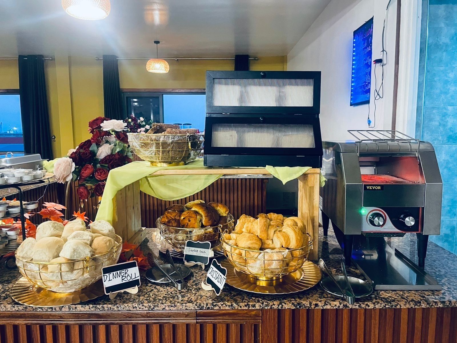 Un bufé con alimentos identificados, como bollos y cruasanes, un horno tostador VEVOR y un arreglo floral decorativo.