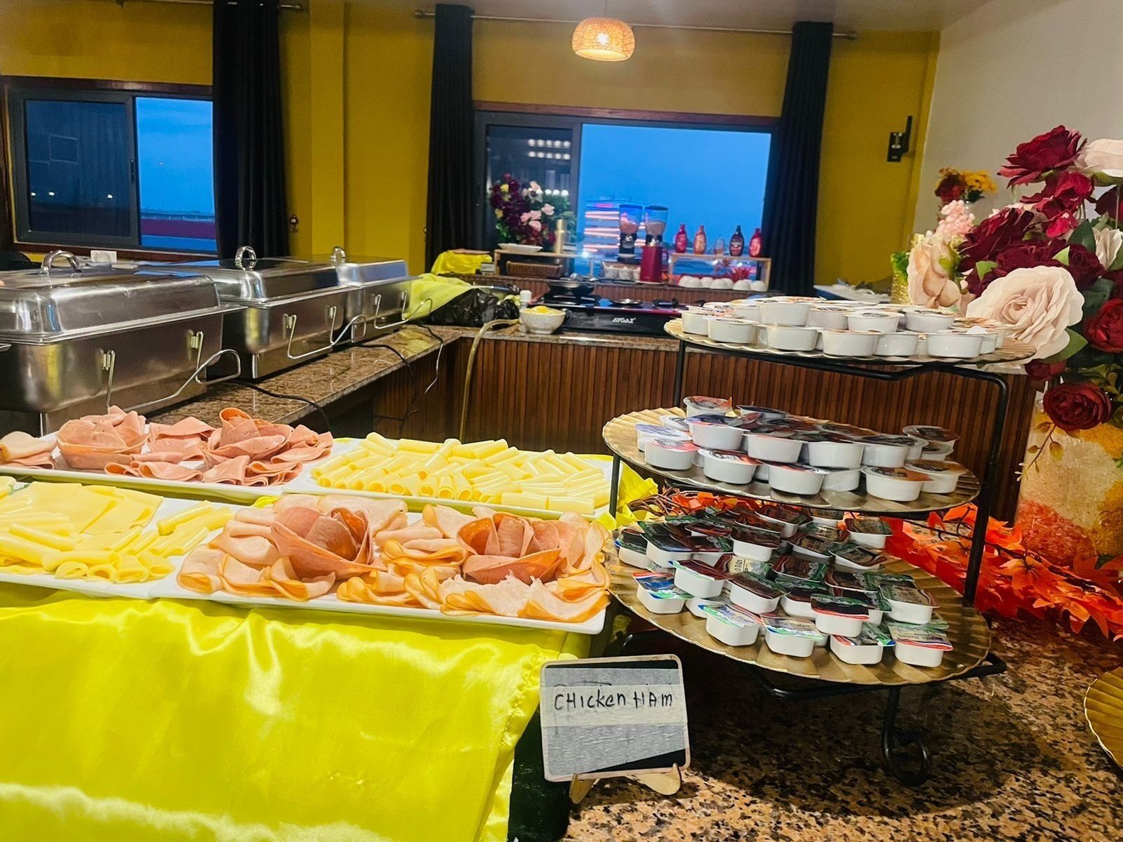 Un bufé con diversos alimentos, incluyendo carnes y quesos, un cartel que dice 'Pollo Jamón', y vistas al mar a través de amplias ventanas.