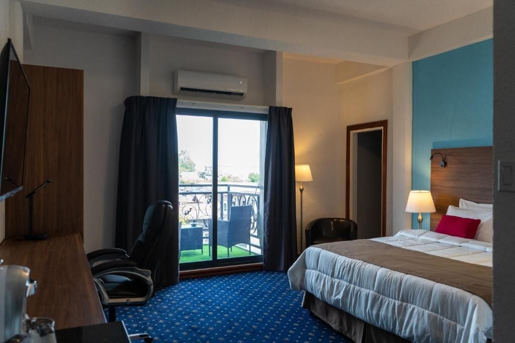 A hotel room with a bed, a desk, a chair, a window with curtains, an air conditioning unit, and a view of the outside.