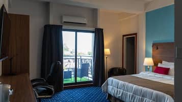 A hotel room with a bed, a desk, a chair, a window with curtains, an air conditioning unit, and a view of the outside.