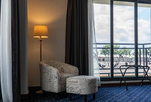 Una habitación con una silla, un reposapiés, una lámpara y vistas a un balcón con barandilla.