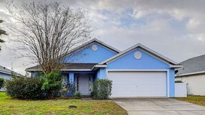 Una casa de un piso con fachada azul, una puerta de garaje blanca y un jardín delantero con un árbol y arbustos.