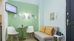Ein Zimmer mit grüner Wand, einem grauen Sofa mit gelben Kissen, zwei weißen Stühlen, einem kleinen runden Tisch mit einer Pflanze, einem an der Wand montierten Fernseher und einer Tür mit Schloss.