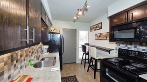 A modern kitchen with dark wood cabinets, a black stove, and a microwave. There is a dining area with a white table and chairs. - Dream House | 2BR Near Galleria (Houston)