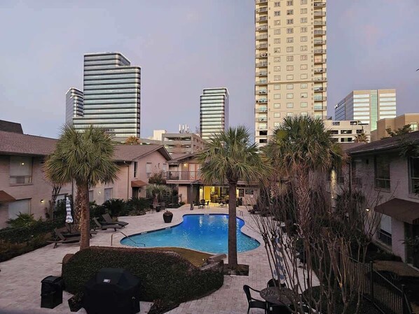 A courtyard with a swimming pool, palm trees, lounge chairs, and high-rise buildings in the background.