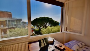A room with a window overlooking a tree and a building. There is a desk with a laptop, a book, and a phone.