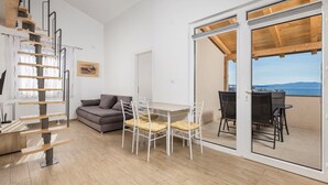 Un interno moderno con una scala in legno, un divano grigio, un tavolo da pranzo con sedie e una porta scorrevole in vetro che dà su un patio esterno con vista.