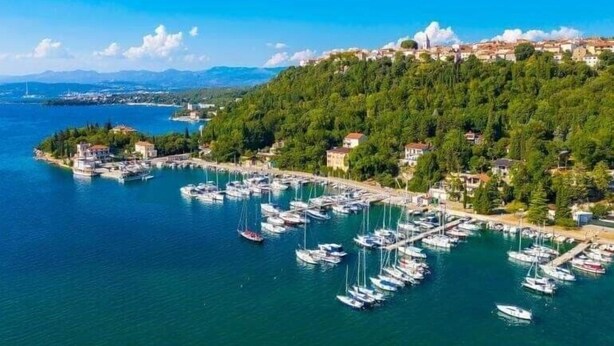 Una cittadina costiera con un porto turistico pieno di imbarcazioni, circondata da vegetazione rigogliosa e un cielo azzurro limpido.