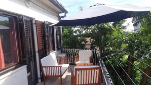Un balcón con sillas y una mesa de madera, una zona cubierta con un paraguas negro y vistas a zonas verdes.