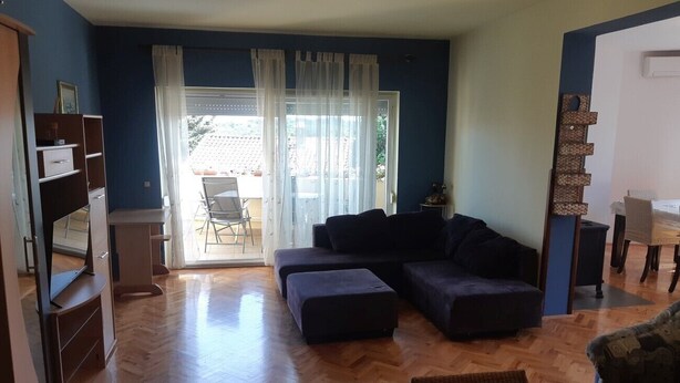 Un salón con sofá, reposapiés y mesa de centro. Se ve una ventana con cortinas, una puerta y una estantería.
