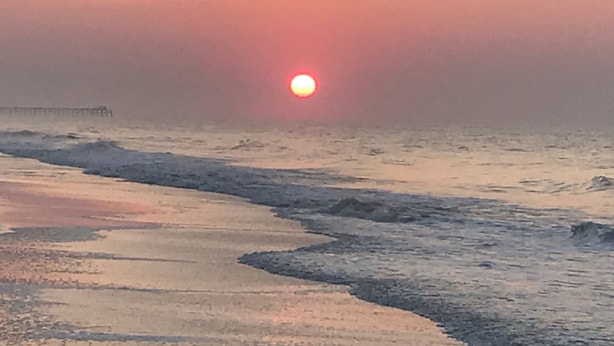 Ein Sonnenuntergang über einem Strand mit Wellen und einem Pier in der Ferne.