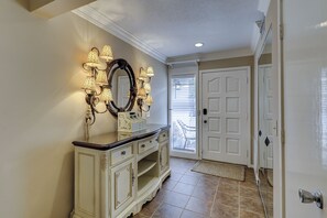 A hallway with a white console table, a large mirror, and wall sconces. There is a door with a window and a doorbell. - Sunset Rentals | Schooner Court 731 (Hilton Head Island)