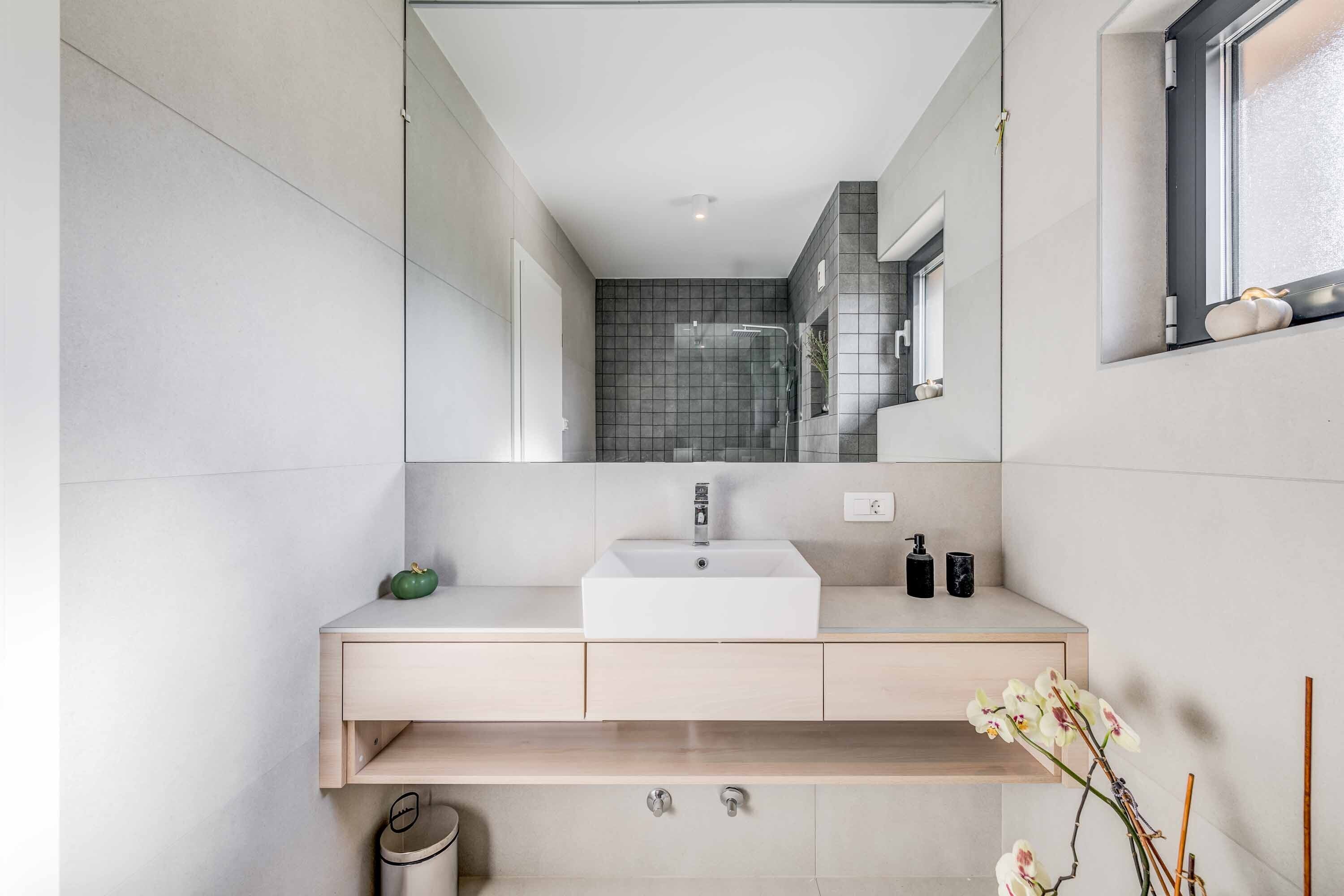 Un bany modern amb un mirall gran, un lavabo blanc i una finestra petita.