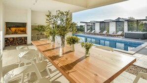 A modern outdoor dining area with a wooden table and white plastic chairs, overlooking a swimming pool with lounge chairs and a hot tub.