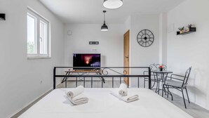 Moderni hotellihuone, jossa on suuri sänky, televisio, ruokailutila pöytineen ja tuoleineen sekä seinällä oleva kello.