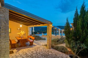 Un patio al aire libre con suelo de piedra, mobiliario de madera y vista a una piscina y montañas al fondo.