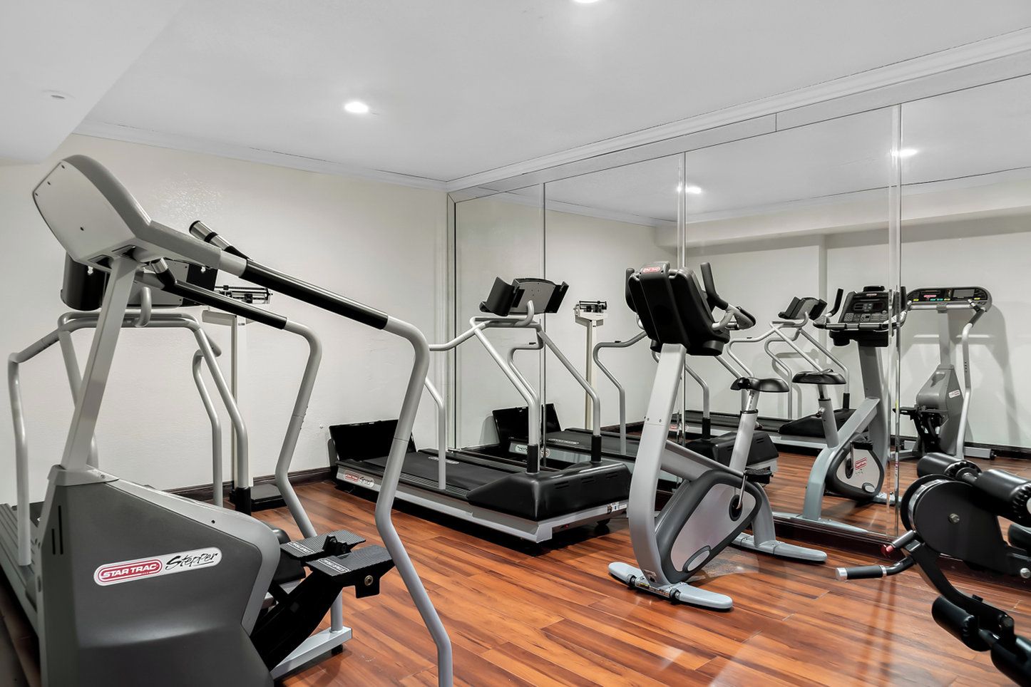 A gym with multiple treadmills and exercise equipment, wooden flooring, and a large mirror on the wall.