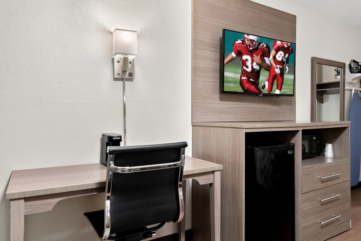 A hotel room with a desk, chair, TV, and a minibar.