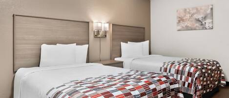 Une chambre d’hôtel avec deux lits, chacun recouvert d’une literie blanche et d’une couverture à carreaux rouges et blancs. Il y a une table de chevet avec une lampe entre les deux lits, et un tableau accroché au mur.